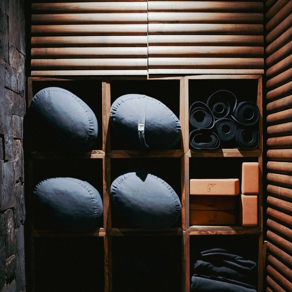 supplies-equipment-square-product Meditation cushions, rolled yoga mats, cork blocks, and folded blankets arranged in warm wooden cubby shelves