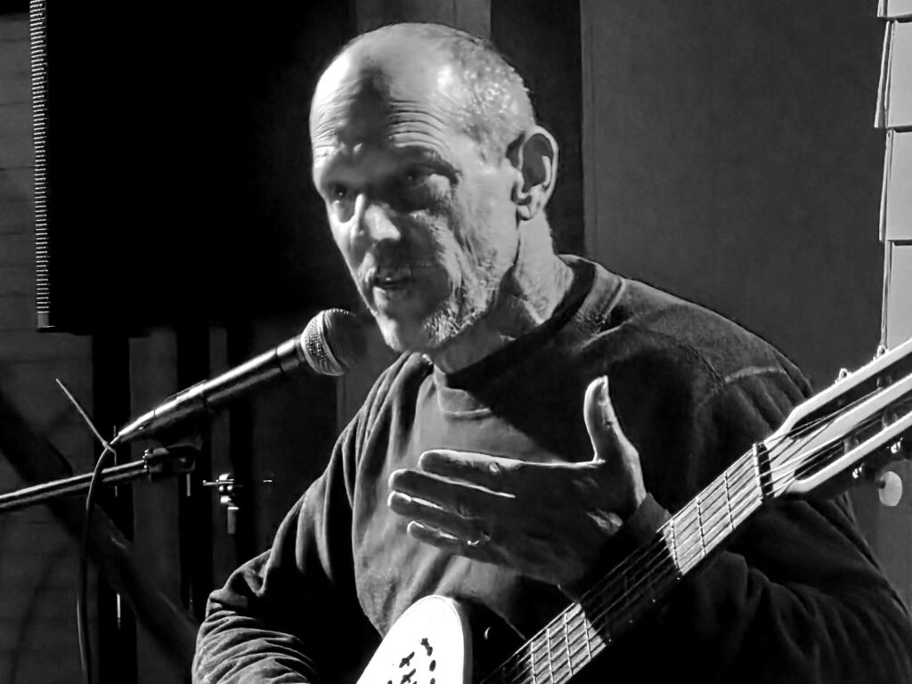 David Kessem with his guitar, speaking into a microphone, hand open at his chest